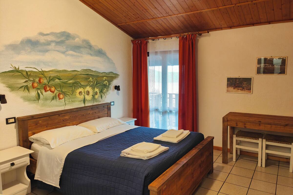 A bedroom with a bed, a wooden headboard, a painting of a landscape, a window with red curtains, and a small table.