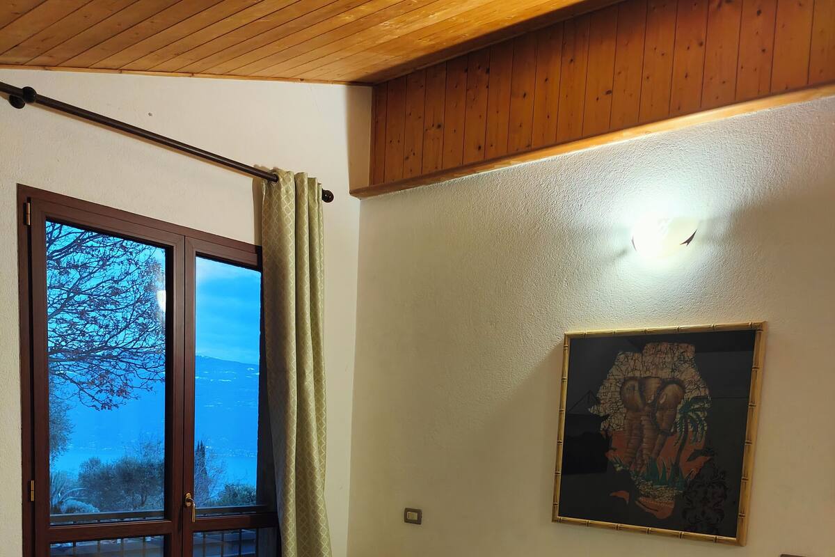 A bedroom with a bed, a window with a view, a painting on the wall, and a wooden ceiling.