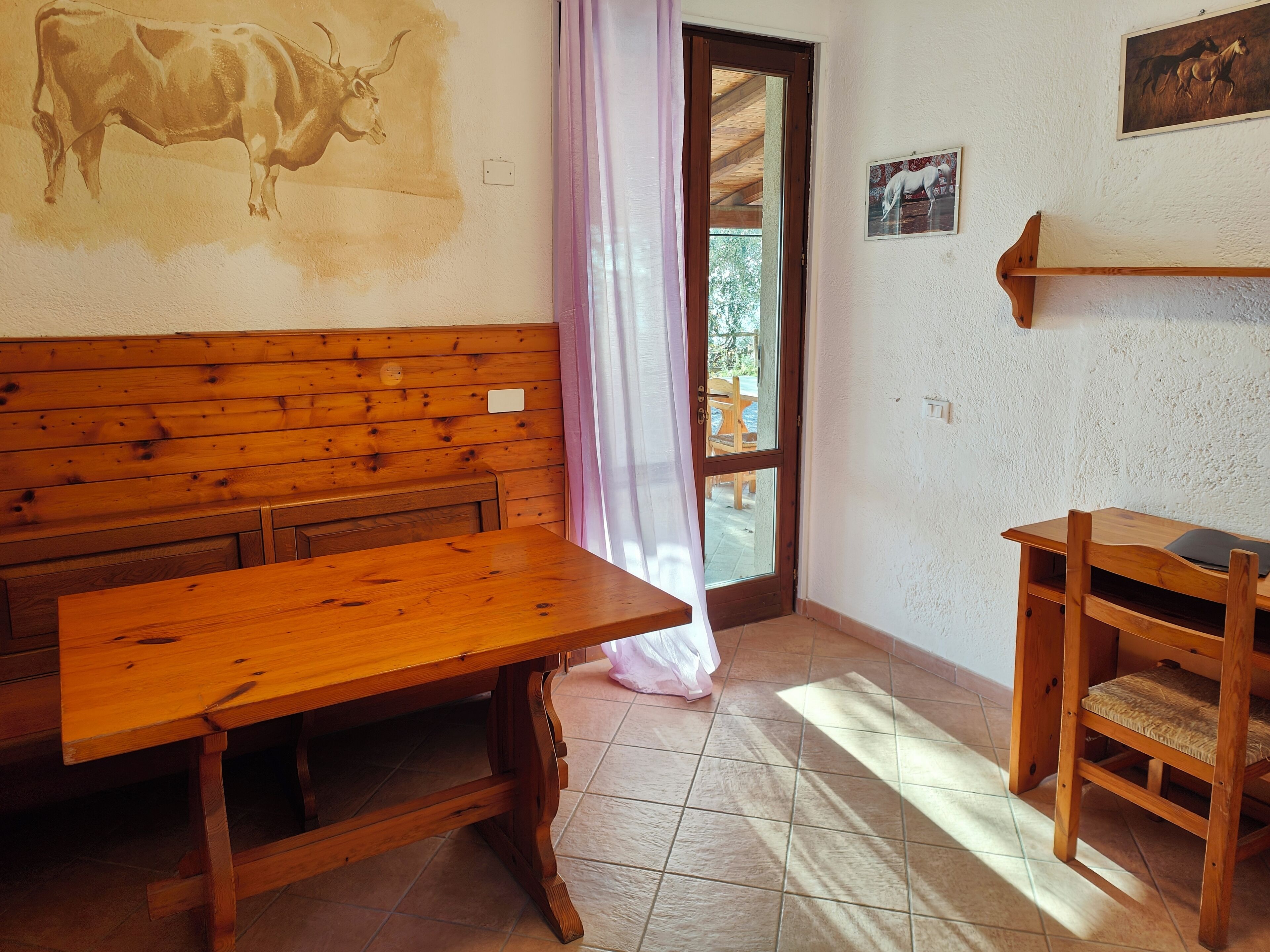 Un banco y una mesa de madera en una habitación de estilo rústico, una puerta que da al exterior y un pequeño estante de madera.