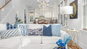 A modern living room with a white sofa, patterned cushions, a chandelier, and a view into the kitchen. - Seagrove Gardens | Mins to the Beach, Santa Rosa Retreat w/ A+ Design (Santa Rosa Beach)