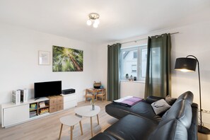Ein modernes Wohnzimmer mit einer schwarzen Ledersofa, einem Flachbildfernseher, einem kleinen Holztisch und einem Bücherregal.