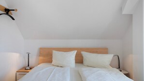 A bedroom with a wooden headboard, two white pillows, a bed with white linens, two bedside tables with lamps, and a wooden shelf on the wall.