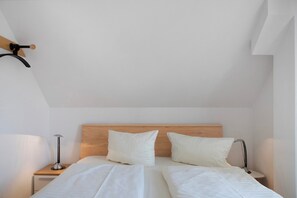 Une chambre avec une tête de lit en bois, deux oreillers blancs, un lit avec du linge de lit blanc, deux tables de chevet avec des lampes et une étagère en bois fixée au mur.