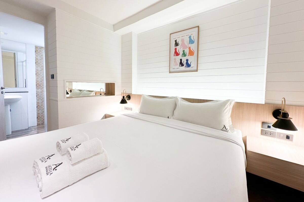 A hotel room with a large bed, a bedside table with a lamp, a wall-mounted picture, and a small window.