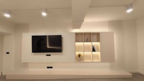 A modern interior with a large flat-screen TV mounted on the wall, a built-in shelving unit with soft lighting, and a minimalist design. - Ascella Pratunam Hotel (Bangkok)