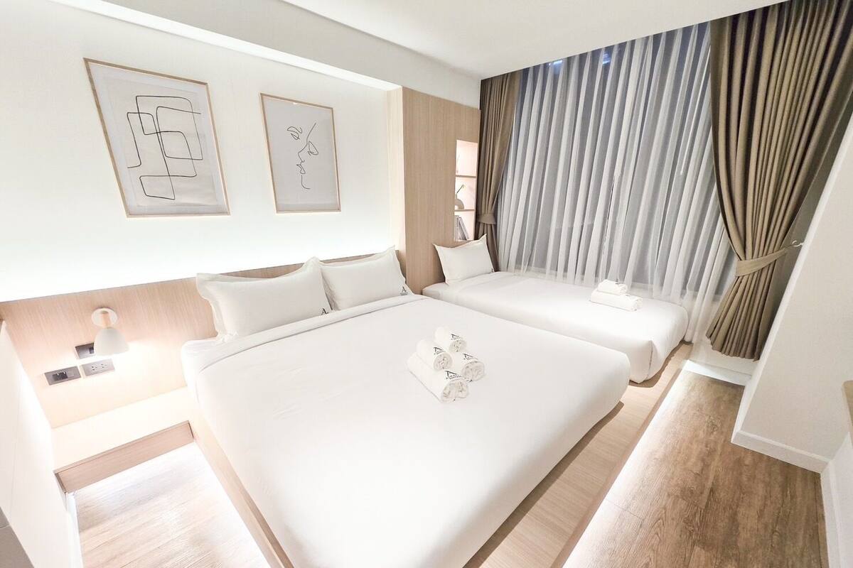 A modern hotel room with a large bed, a smaller bed, and a desk. There are two framed artworks on the wall, a window with curtains, and a wooden floor.
