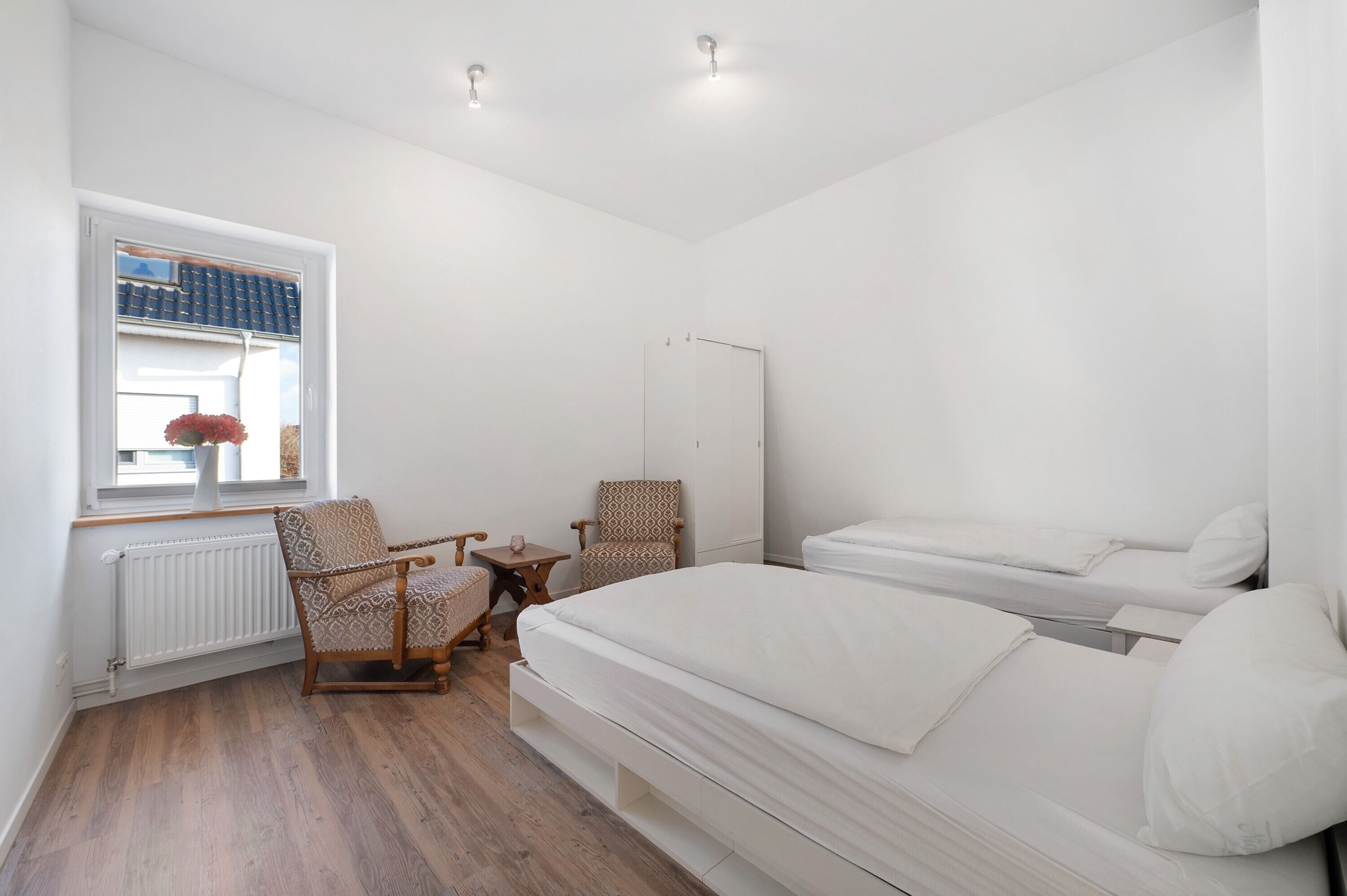 Une chambre à coucher comprenant un lit, deux chaises, une petite table, un radiateur et une fenêtre donnant sur un immeuble et des fleurs.