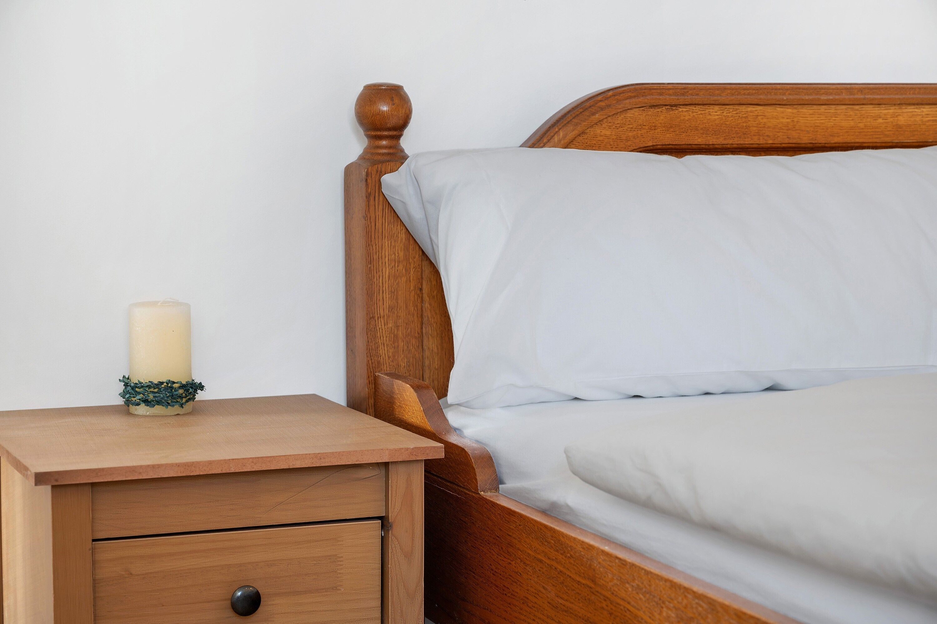 Een houten nachtkastje met een lade, een brandende kaars en een bed met wit beddengoed.
