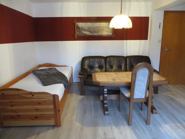 A room with a bed, a sofa, a dining table with chairs, a hanging light fixture, and a door.