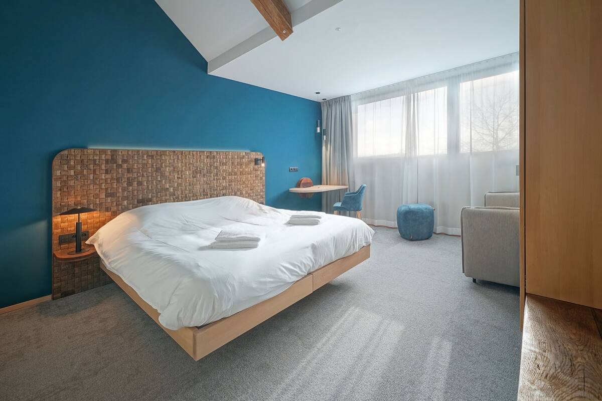 A modern hotel room with a large bed, a small desk, a chair, and a view of the outdoors through large windows.