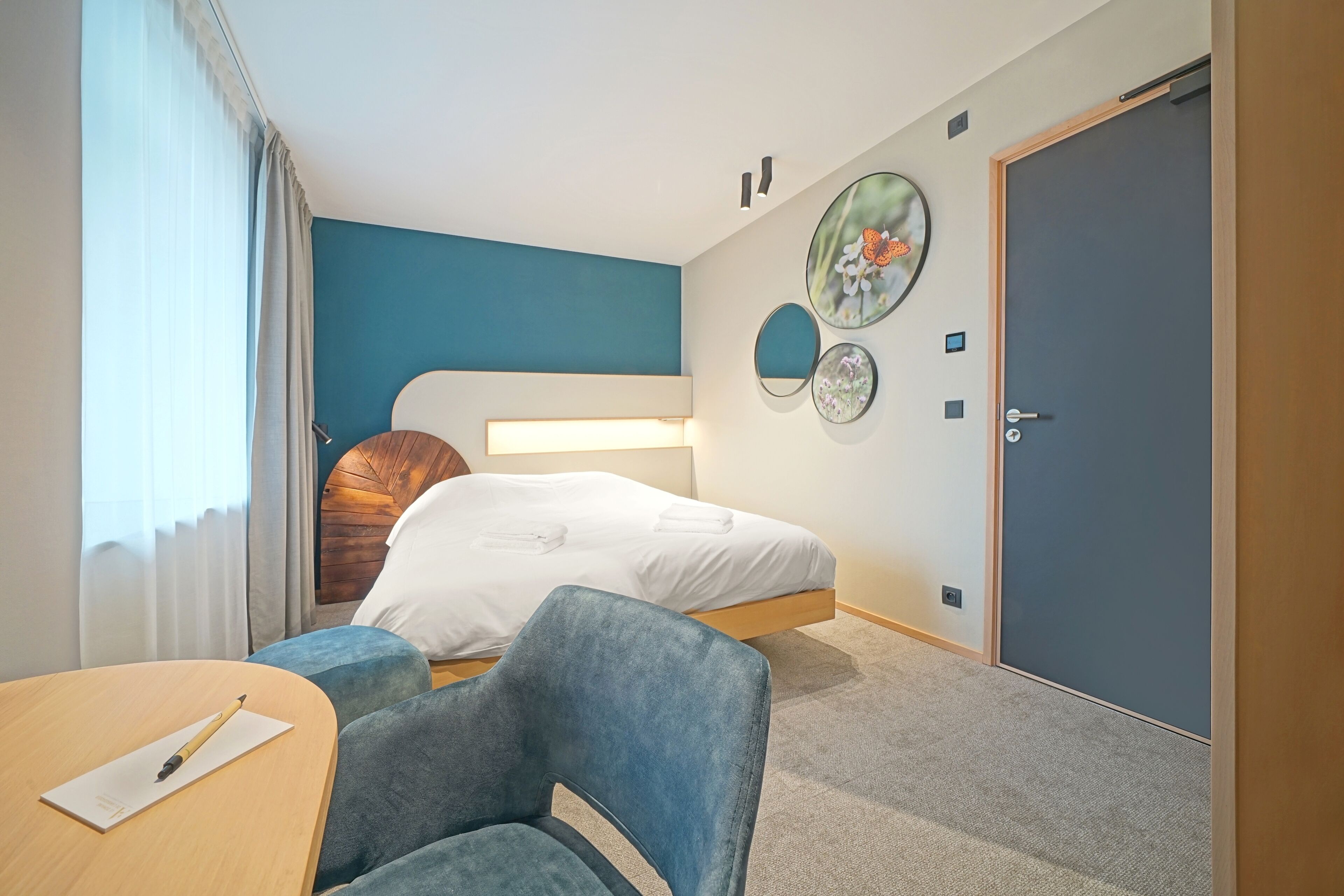A hotel room with a bed, a desk with a pen, a chair, a door, and wall decorations.