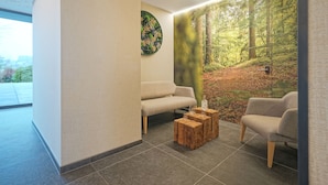 Un espace d’attente moderne, aménagé avec un canapé, deux fauteuils et une table basse en bois. Une grande fresque murale représente une scène de forêt.