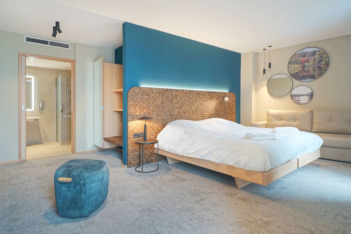 A modern bedroom with a bed, a blue ottoman, a small table, and a wall-mounted mirror.