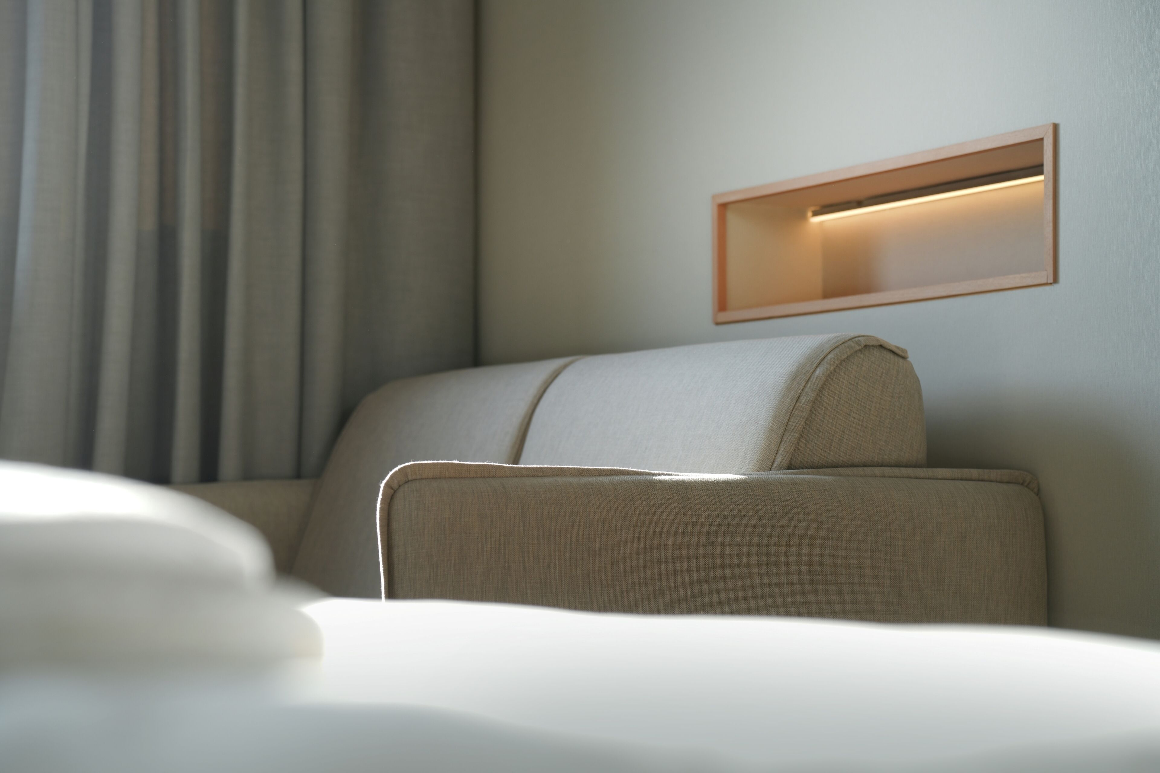 A modern hotel room with a bed, a headboard with built-in lighting, and a large window with curtains.