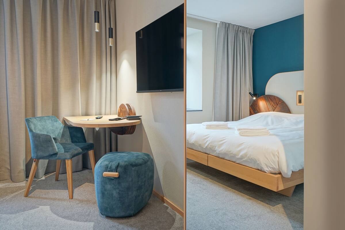A hotel room with a bed, a desk with a chair, a TV, and a small ottoman.