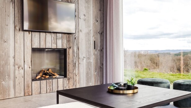 A modern interior with a wooden wall, a fireplace, a flat-screen TV, and a dining area with a table and ottomans. - Modern Maison Near Somme-leuze (Somme-Leuze)