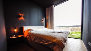 A modern bedroom with a large bed, a bedside table with a lamp, and a view of a grassy landscape through a large window. - Modern Maison Near Somme-leuze (Somme-Leuze)