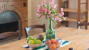 Una mesa de madera con un bol de manzanas verdes y un bol de melocotones rojos y amarillos, un jarrón verde con flores rosadas, y una chimenea de ladrillo al fondo.