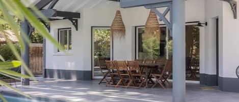 A covered outdoor dining area with a pool, wooden chairs, and hanging light fixtures.