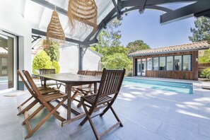 Eine Terrasse im Freien mit einem runden Holztisch und Stühlen, einem Pool und einem modernen Haus im Hintergrund.