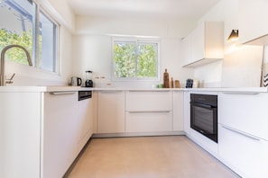 Eine moderne Küche mit weißen Schränken, einem eingebauten Backofen, einem Fenster mit Blick auf Bäume und einer Arbeitsfläche mit einem Wasserkocher und anderen Gegenständen.