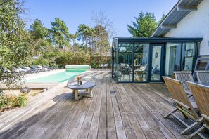 Une terrasse en bois avec un espace patio fermé par des baies vitrées, une piscine et des sièges d’extérieur.