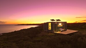 A small, modern cabin with a glass door and solar panels, situated on a grassy hill overlooking a body of water during sunset. - Poolside Ocean View Pod (Marion Bay)