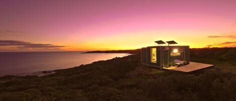 A small, modern cabin with a glass door and solar panels, situated on a grassy hill overlooking a body of water during sunset.