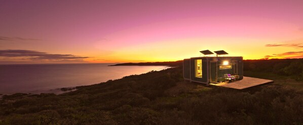 A small, modern cabin with a glass door and solar panels, situated on a grassy hill overlooking a body of water during sunset.
