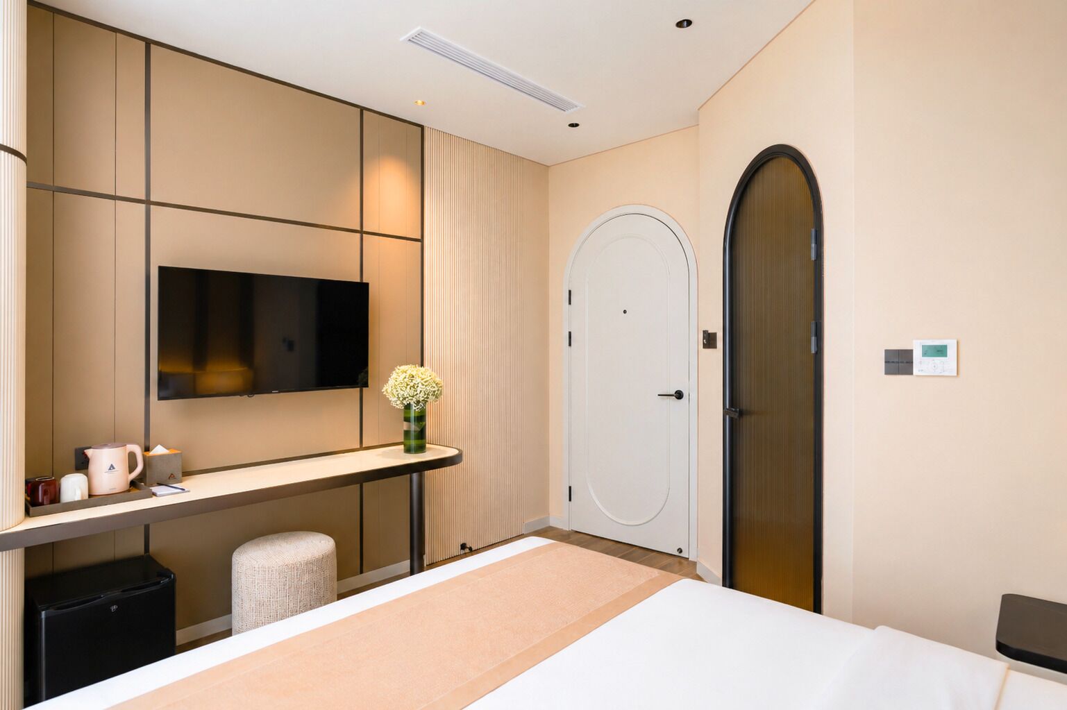 A hotel room with a bed, a TV mounted on the wall, a desk with a vase of flowers, a trash bin, and a door.