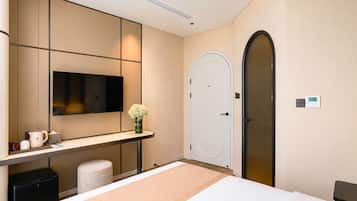 A hotel room with a bed, a TV mounted on the wall, a desk with a vase of flowers, a trash bin, and a door.