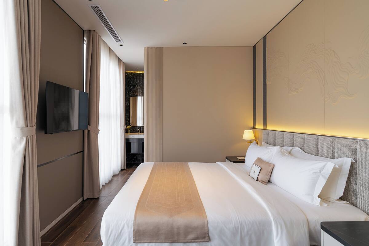 A hotel room with a large bed, a flat-screen TV mounted on the wall, a nightstand with a lamp, and a view of the bathroom and hallway.