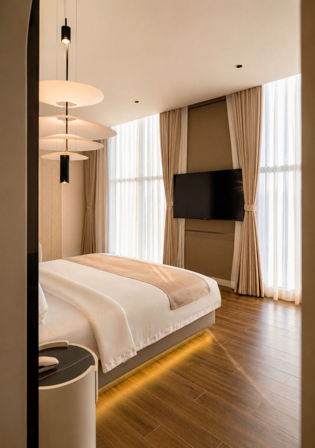 A modern bedroom with a large bed, a flat-screen TV mounted on the wall, and a large window with curtains.