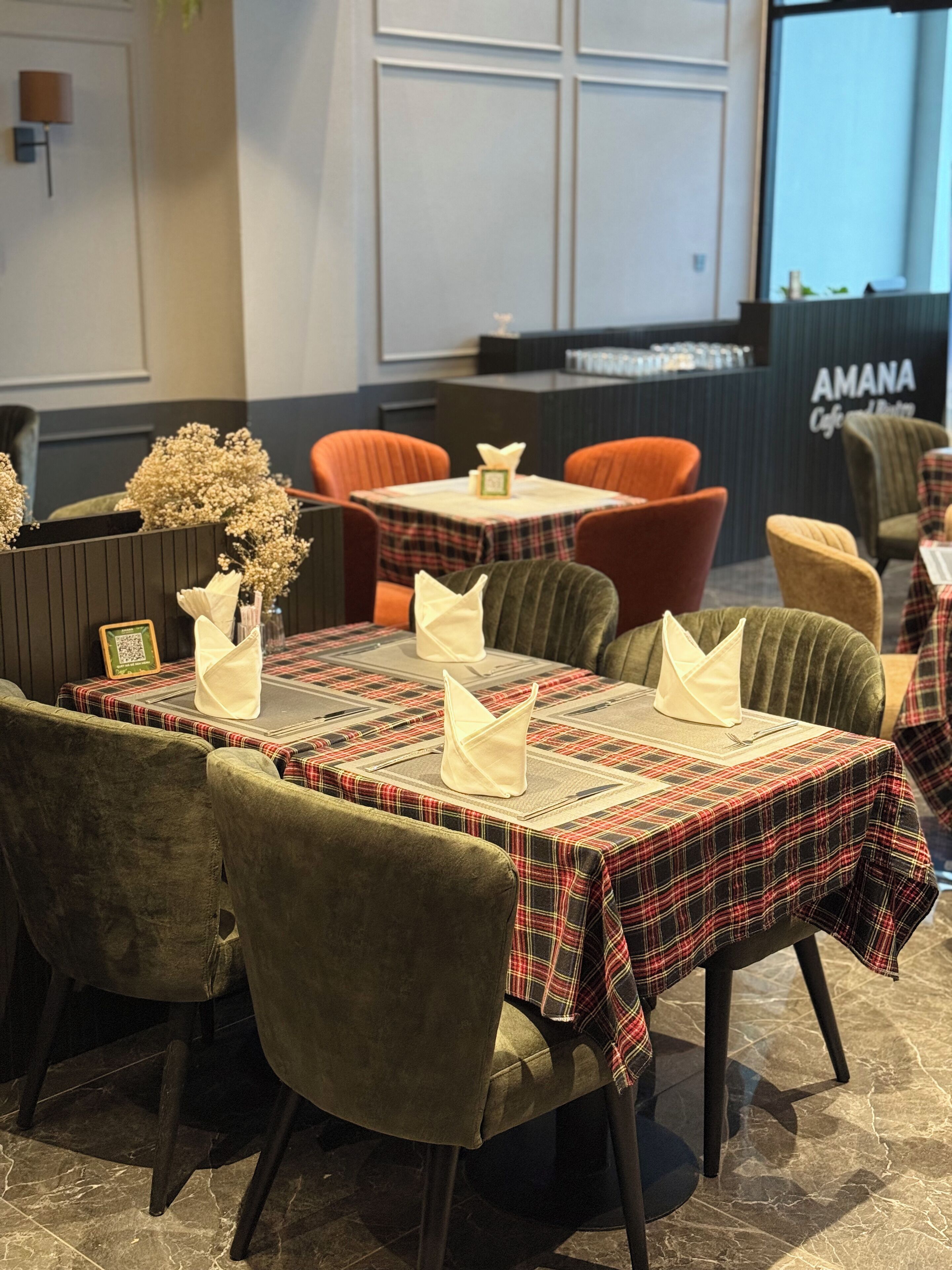 A restaurant with plaid tablecloths, green upholstered chairs, and a counter with a sign reading "AMANA Cafe & Bar".