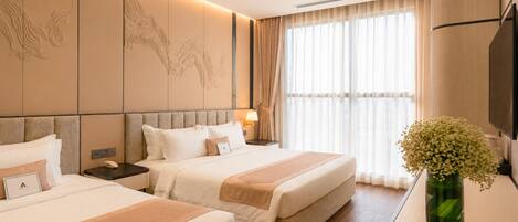 Une chambre d’hôtel avec deux lits, une grande fenêtre avec des rideaux légers, un téléviseur fixé au mur et un vase rempli de fleurs placé sur une table d’appoint.