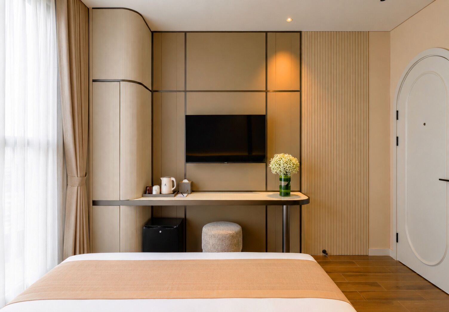 A modern hotel room with a bed, a bedside table, a TV, and a small stool.