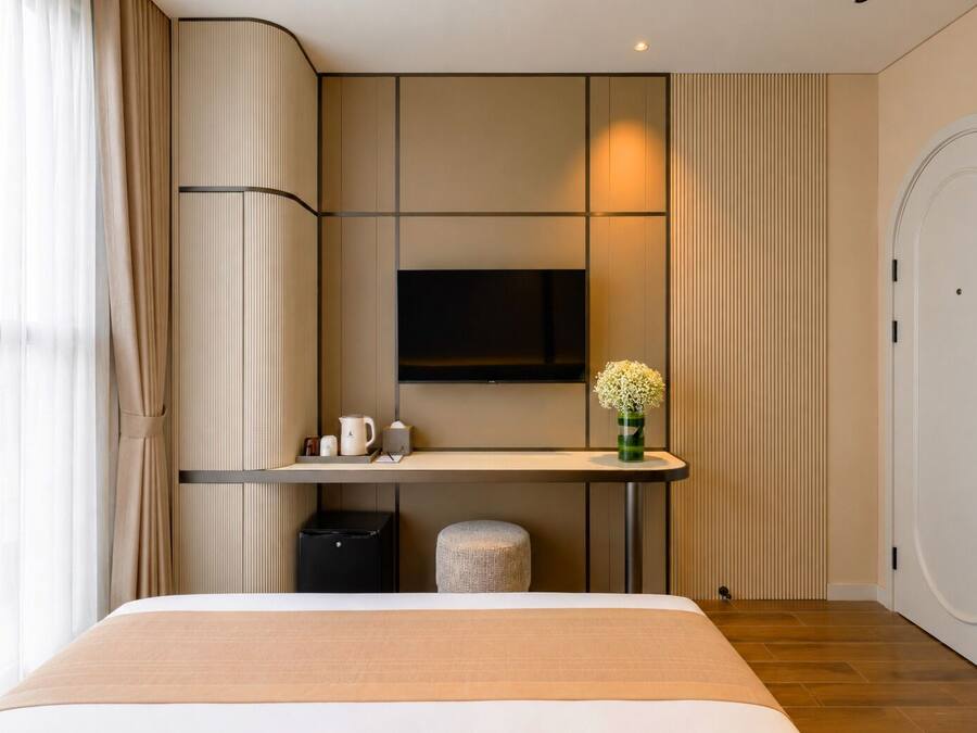 A modern hotel room with a bed, a bedside table, a TV, and a small stool.