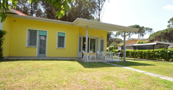 A yellow building with a covered patio area, white chairs, and a small garden.