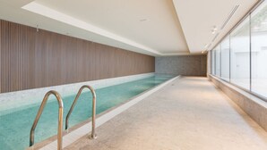 A swimming pool with a tiled floor and metal handrails, a wooden wall, and a large window on one side.
