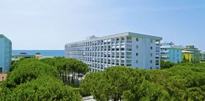 A modern hotel with a grid-like facade, surrounded by lush greenery and a clear view of the sea. - Seafront studio in Bibione (Bibione)