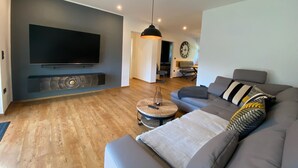 A modern living room with a large flat-screen TV mounted on the wall, a grey sofa with patterned cushions, a round coffee table, and a dining area in the background. - Vacation home Bimmelbahn track 2 (Olbersdorf)