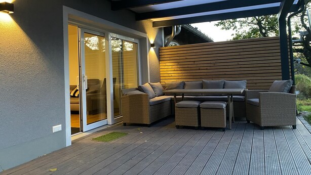An outdoor patio area with a covered seating arrangement, a sliding glass door leading to an interior space, and a wooden deck. - Vacation home Bimmelbahn track 2 (Olbersdorf)