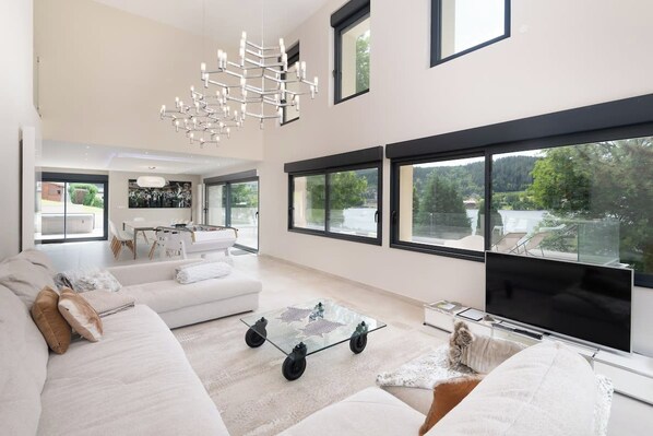 A spacious living room with a large flat-screen TV, a glass coffee table, and a modern chandelier.