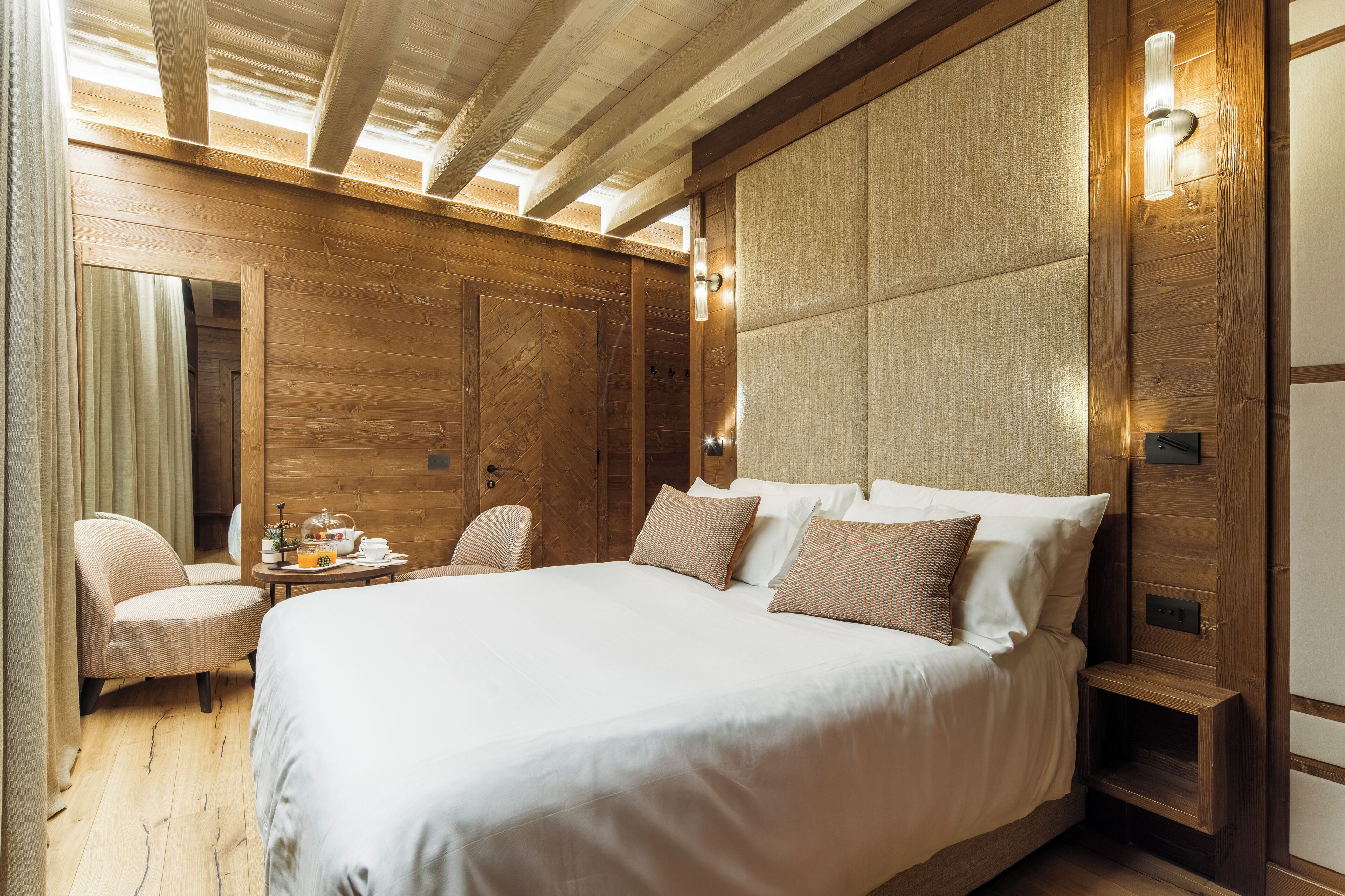 A large bed with white linens, a wooden headboard, and two bedside lamps. There is a small table with a lamp and a chair to the left of the bed.