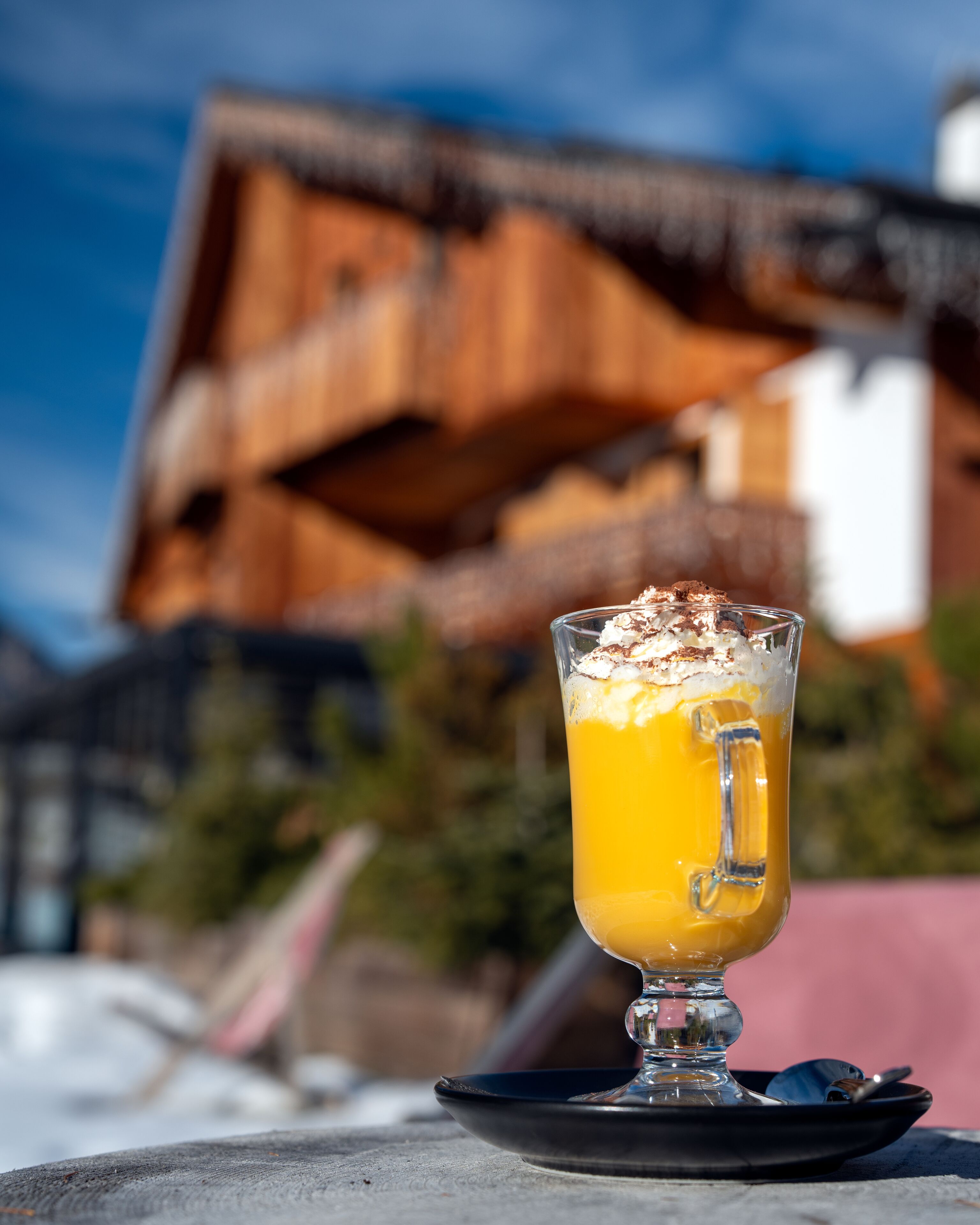 Um copo de suco de laranja com chantilly sobre uma mesa com um apoio para copos preto, em frente a um prédio com telhado inclinado.
