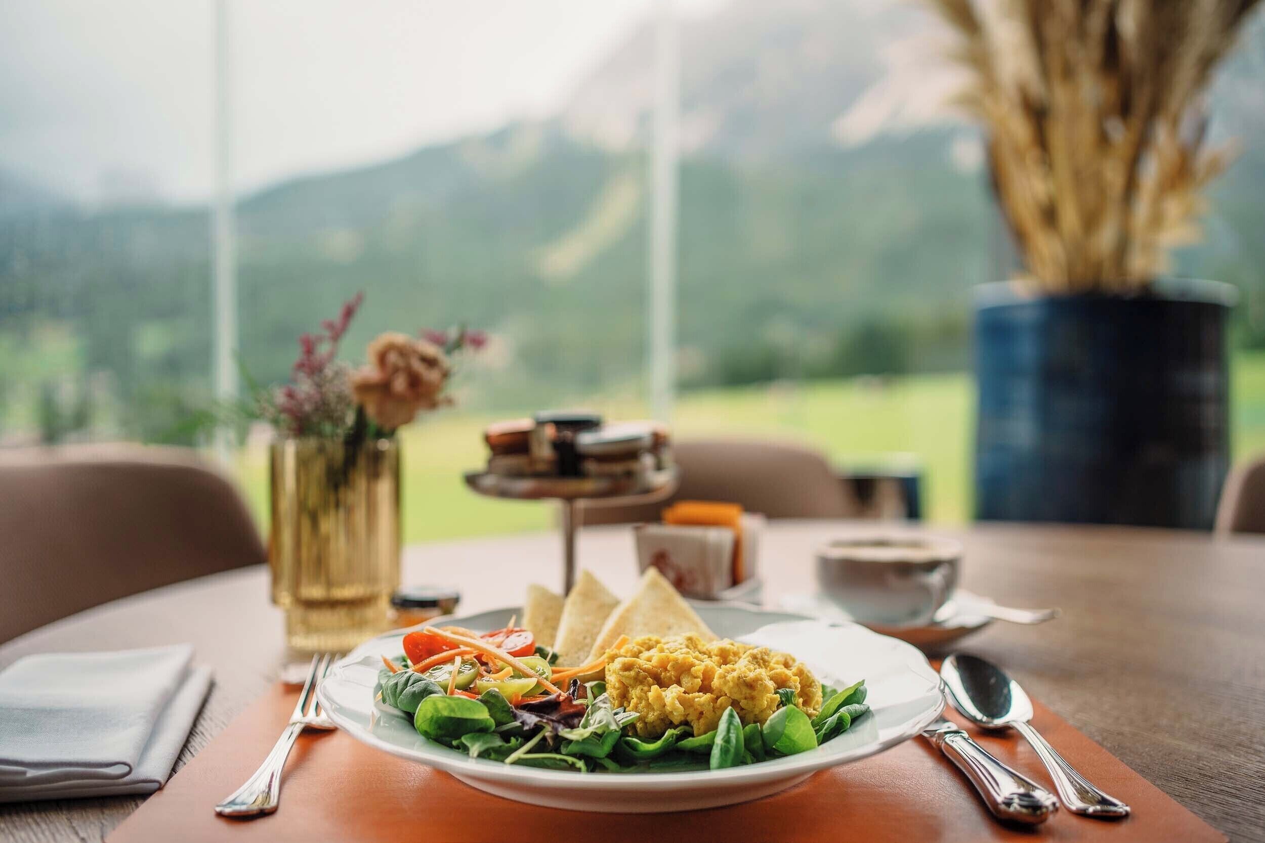 Un plato de huevos revueltos, tostadas y ensalada sobre una mesa con vistas a las montañas.