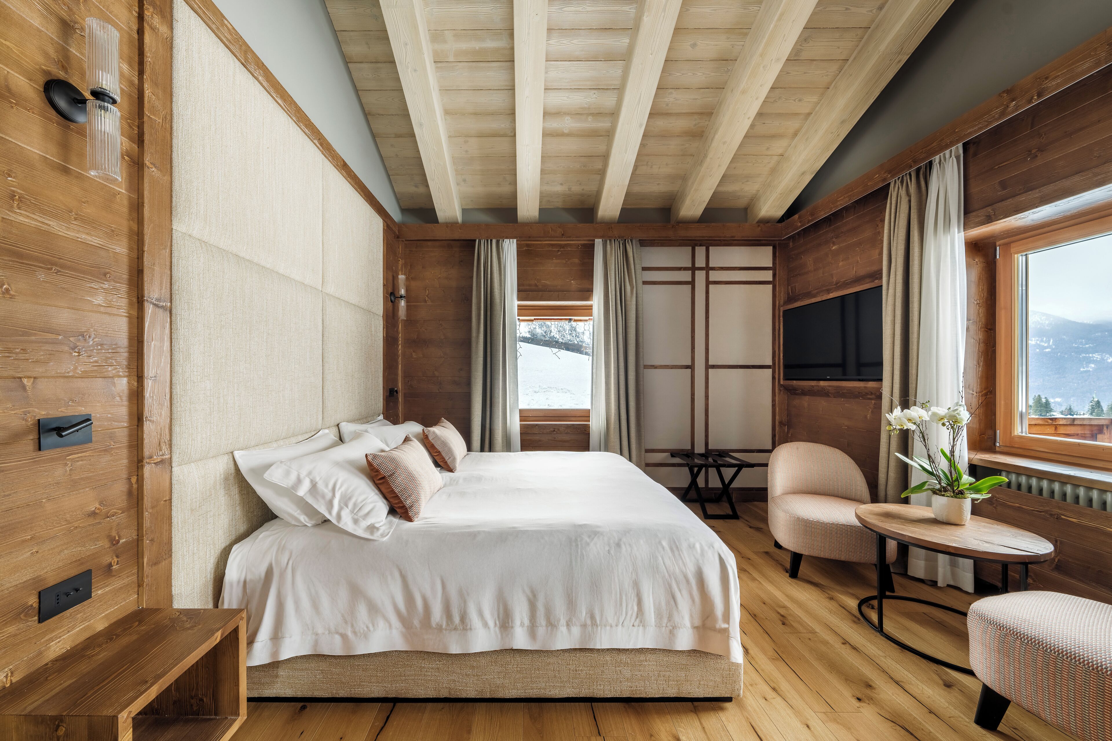 Una habitación de hotel moderna con techo de madera, una cama grande con ropa blanca, una zona de estar con una mesa y vistas a la montaña a través de la ventana.