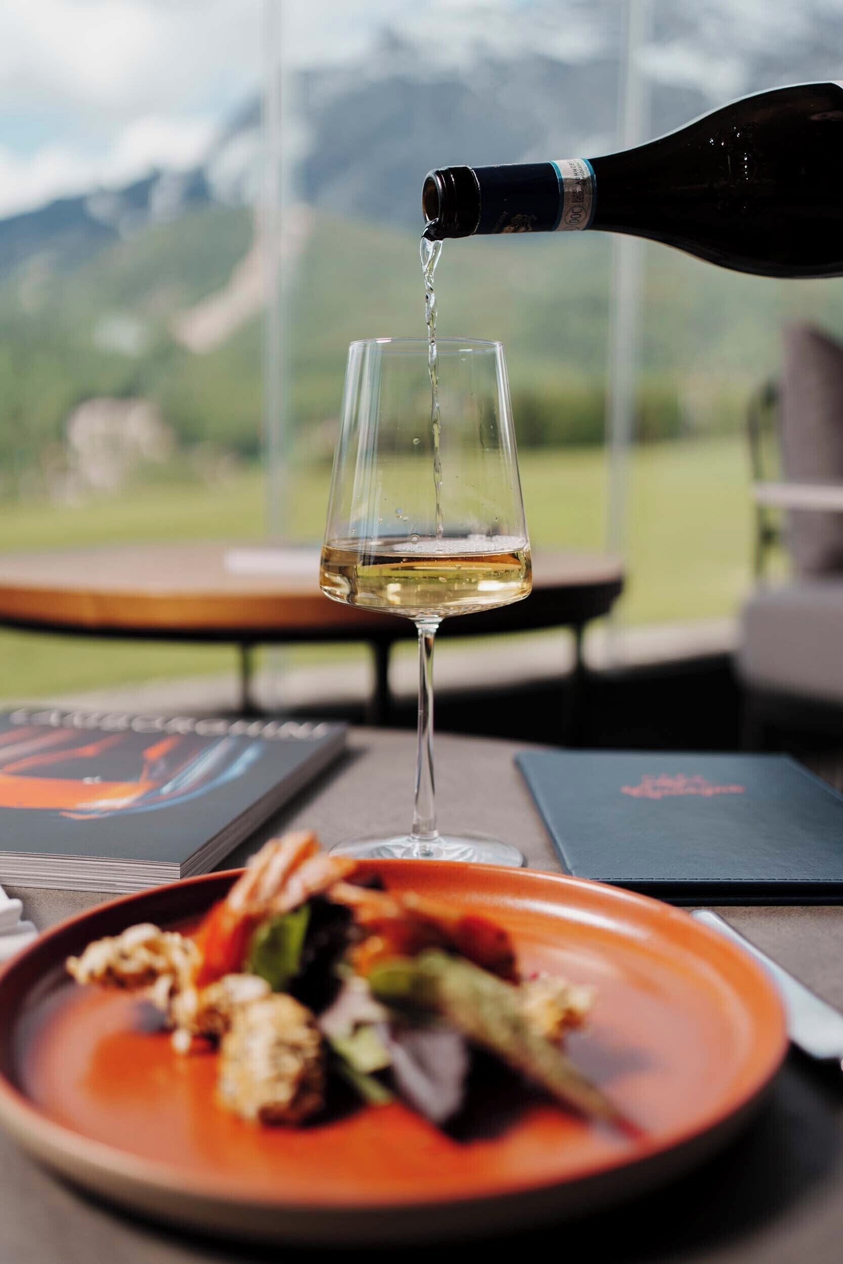 Un verre de vin blanc est versé d’une bouteille, une assiette de nourriture et un menu sont posés sur une table, avec une vue sur les montagnes en arrière-plan.