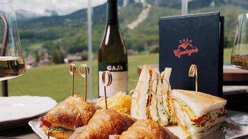 A table with croissants, sandwiches, a wine bottle, and a menu with a mountain view in the background.