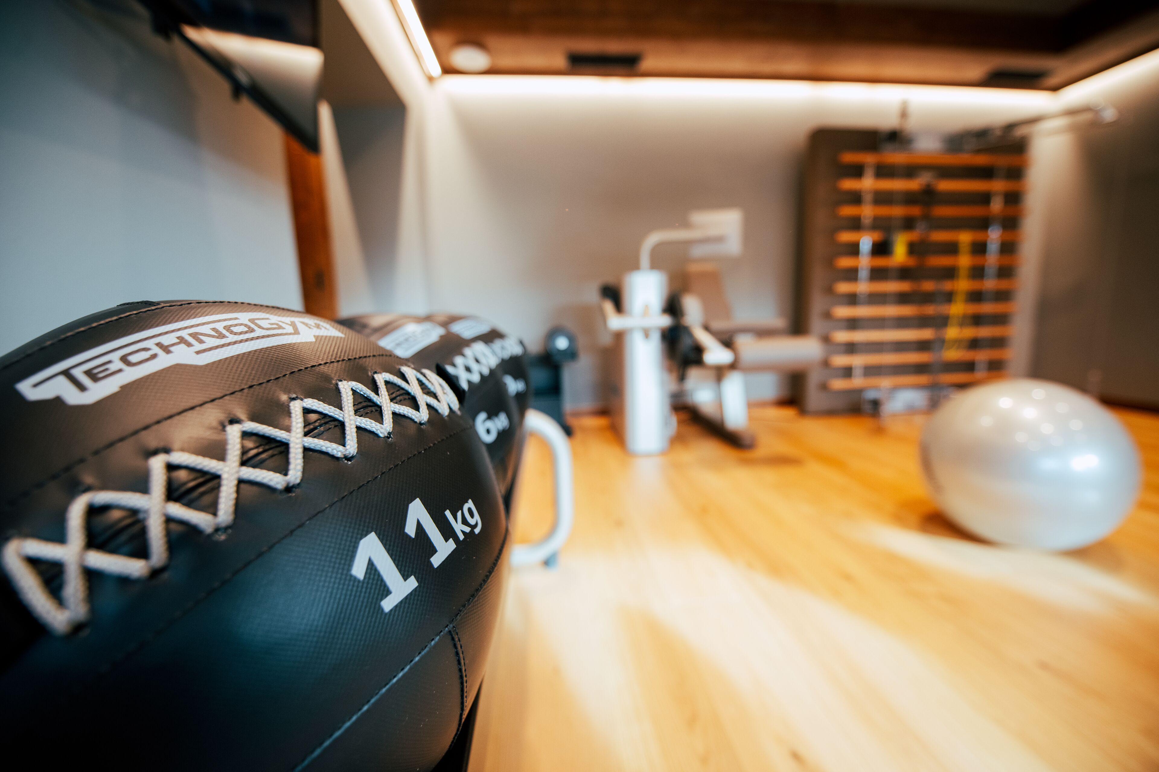 A close-up of a sports ball with 'TECHNOGYM' and '11 kg' visible, a white exercise machine, and a wooden floor in a room with a slanted ceiling.